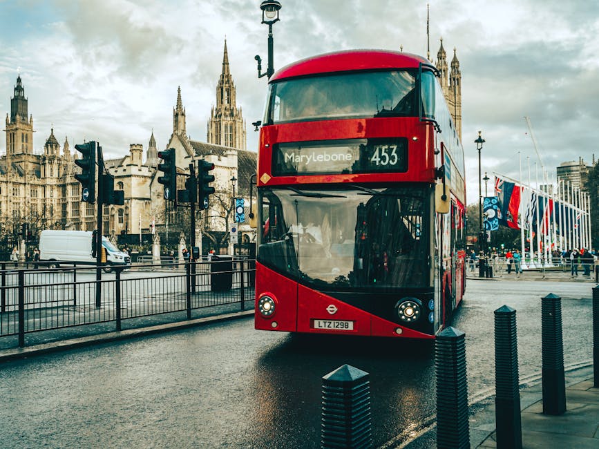 A red double-decker bus with a modern, streamlined design and large, curved front windows is approaching a bus stop on a wet urban street. The bus displays route number 453, serving the Marylebone area, and is branded with the London Transport logo on the front. Behind the bus, there are historic Gothic-style buildings with pointed spires and intricate stonework, alongside a lamppost and a row of flags fluttering in the breeze. To the left, there is a white delivery van traveling along the road, which is separated from the bus stop by a black metal railing. The street surface appears slick from recent rain, with reflections of the bus and surrounding environment visible on the wet pavement. On the right, black waist-high bollards are positioned along the sidewalk, and a few pedestrians are visible in the background, some walking near the flags and some waiting nearby. This scene captures aspects of urban transportation and infrastructure, consistent with private rubbish removal services like those offered by Rubbish Removal Marylebone, which support alternative waste handling beyond public collection systems.