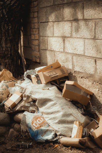 A collection of discarded cardboard boxes, some flattened and others partially assembled, along with a plastic bag containing unknown waste items, all piled against a rough, weathered brick wall outdoors. The boxes are light brown with printed labels and barcodes, made of corrugated cardboard with visible ridges, and exhibit creases and torn edges. Surrounding the waste are small rocks and dirt on the ground, with a large dark tree trunk visible on the left side of the scene. The background wall is constructed of beige bricks with a slightly textured surface, and the overall environment appears to be an alleyway or outdoor storage area, suggesting a context of private waste disposal or rubbish collection. This scene is typical of independent rubbish removal efforts, where waste materials are temporarily stored prior to collection or disposal, relevant to the services offered by Rubbish Removal Marylebone in managing excess rubbish outside of regular council collection schedules.