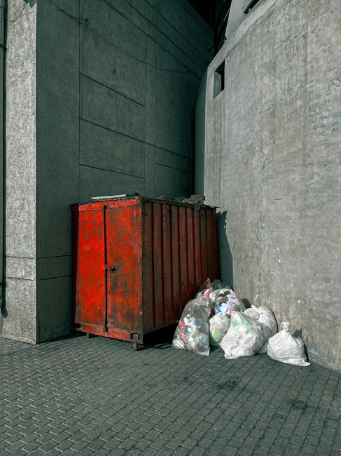 A large, rusty red metal dumpster with vertical ridges and a slightly weathered surface is positioned against a dark green wall on the left side and a light grey concrete wall on the right side. The dumpster is situated on a paved surface composed of small, interlocking dark grey bricks, with some debris visible on top. Several clear plastic rubbish bags filled with waste, including visible contents like plastic bottles and paper, are piled around the base of the dumpster, some leaning against it. The environment appears to be an alleyway or side street area, with the space confined by the two walls. Natural light illuminates the scene, casting soft shadows, and the overall setting suggests a location for private waste disposal, possibly an alternative rubbish collection point handled by a service such as Rubbish Removal Marylebone. The scene's neutral tones and the presence of waste suggest an outdoor rubbish removal context typical of independent collection or on-site clearance services.