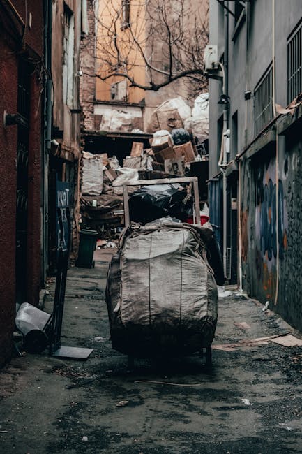 A narrow urban alleyway filled with waste and debris, with a large black and grey bag in the foreground containing disposed materials such as cardboard, plastic, and paper. Behind the bag, there are numerous stacks of cardboard boxes, black garbage bags, and other rubbish piled high against a dilapidated wall at the end of the alley. The surrounding buildings feature graffiti on the walls, with one side showing a grey wall with protective metal bars and the other side having a brick and plaster façade. Bare, leafless tree branches extend overhead against a cloudy sky, creating a stark atmosphere. The ground is uneven and stained, with scattered trash and dirt, indicating a private or alternative waste removal scenario often handled by specialist services like Rubbish Removal Marylebone, who manage bulky waste and debris clearance in such urban environments.