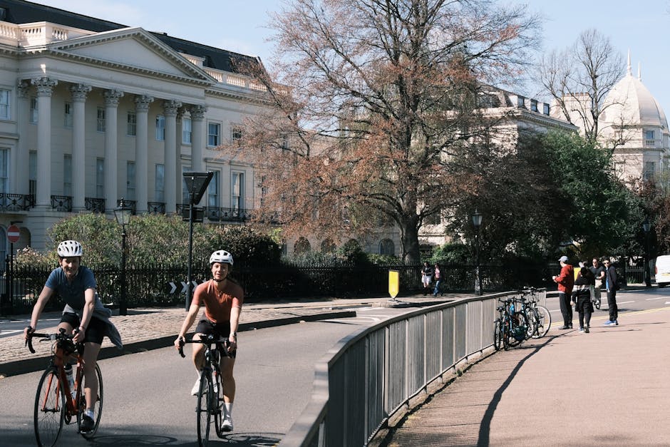 A street scene in an urban area showing two cyclists, a man and a woman, riding on the road while wearing protective helmets; the man is dressed in a dark T-shirt and shorts, and the woman in a reddish-brown top and white shoes. On the right side, there is a paved sidewalk with three bicycles secured to a metal railing. Several pedestrians are walking or standing, with some engaging in conversation. The background features a stately white building with classical architectural details, including tall columns and decorative moldings, partially obscured by large, leafless trees with sprawling branches. The scene is illuminated by natural daylight, casting mild shadows, and the overall environment appears calm and orderly, reflecting a typical cityscape that could be associated with public or private transportation options, fitting the theme of alternative waste management and rubbish removal services such as those offered by Rubbish Removal Marylebone.