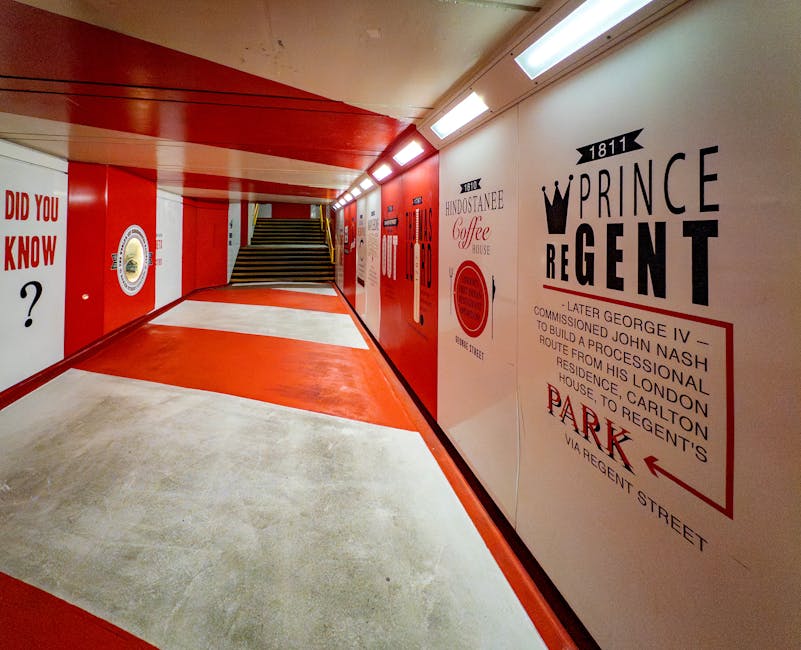 A brightly lit underground passageway with a predominantly red and white colour scheme, showing a smooth, polished concrete floor with red painted sections and white borders. The ceiling features a series of rectangular fluorescent lights, providing uniform illumination across the space. On the right-hand wall, there is a large graphic sign with black text and a crown symbol, displaying historical information about the Prince Regent and a directional arrow pointing left towards a parking area on Regent Street. The left wall features a circular emblem with a gold border and an unclear central design, alongside text and other illustrative graphics. A flight of wide, dark steps with wooden or dark metal risers and treads leads upward at the far end of the corridor. The environment suggests an official or institutional setting, possibly a pedestrian underpass or access corridor associated with a historic venue or public space, with no visible clutter or debris. The space appears clean, well-maintained, and designed for safe, independent movement, aligning with private or alternative rubbish disposal concepts that Rubbish Removal Marylebone might support in waste management services.