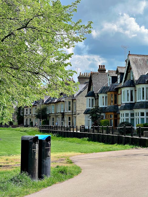 A row of Victorian-style terraced houses with bay windows and slate roofs lines a residential street. In the foreground, two black cylindrical rubbish bins with blue and green lids are positioned near a concrete pathway. To the left, a large tree with fresh green leaves extends its branches over the scene, casting partial shadows on the grassy area and pavement below. The background features a partly cloudy sky with patches of blue visible through the white clouds. The environment suggests a peaceful urban neighborhood, where local waste collection might be supplemented by private rubbish removal services such as those offered by Rubbish Removal Marylebone, indicating a typical scene of communal and private waste management efforts in a well-maintained residential area.