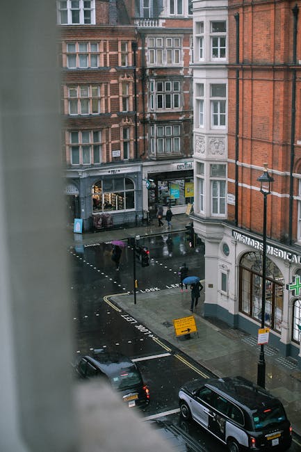 View of a wet city street on a rainy day, taken from a high vantage point through a window frame from a building. The street is lined with multi-storey redbrick and white-painted buildings with large bay windows. Pedestrians, some holding umbrellas, walk along the sidewalk and cross at a pedestrian crossing. Several vehicles, including a parked black SUV and a grey car, are present on the road. A small white sign with black text is placed on the roadway, indicating temporary traffic control or parking restrictions. Street lamps line the pavement, and there is a shop with a dark awning and brightly colored signs visible on the ground floor of one building. The scene captures the typical urban environment, and the wet surfaces reflect the overcast sky, creating a reflective sheen on the pavement. This image is relevant to private waste collection or on-site clearance services like those provided by Rubbish Removal Marylebone, illustrating the urban context where such services operate.