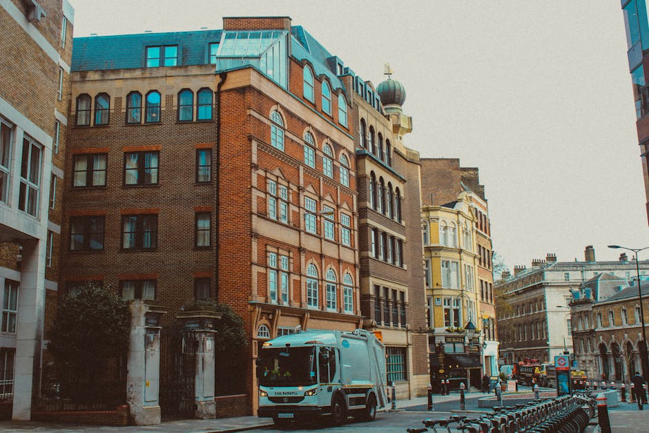 A light blue waste collection vehicle, branded with the company's logo, is parked on a city street alongside multi-story buildings with varied architectural styles. Situated in front of an ornate residential building with brickwork, arched windows, and decorative stone details, the vehicle appears to be preparing for rubbish removal or on-site clearance. To its left, a dark wrought-iron fence and stone pillars mark an entrance to a property, partially obscured by trees. The street is lined with a row of black bike racks, some of which are empty, while the background shows additional historic buildings with intricate facades and a mix of modern and traditional architecture. The environment suggests a busy, urban area consistent with private waste handling operations, highlighting the type of alternative rubbish collection services offered by Rubbish Removal Marylebone. The scene is evenly lit, with no harsh shadows, reflecting typical daytime lighting suitable for efficient rubbish removal efforts within a city context.