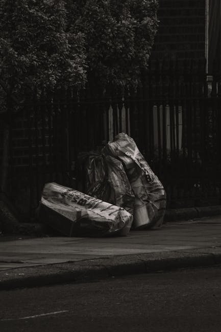 Three large black garbage bags filled with waste material are placed on the sidewalk near a black metal fence and a brick wall. The bags are made of thick plastic and appear to contain household or garden refuse, with some partially bulging and showing creases and folds on their surfaces. They are positioned in a loose cluster, with one bag leaning against the fence and the other two lying flat on the pavement. The scene is outdoors, with an area of greenery visible behind the fence, and natural light illuminating the scene, creating subtle shadows on the bags and the ground. The image conveys an outdoor rubbish collection setting, typical of private waste removal services offered by companies like Rubbish Removal Marylebone, and indicates a situation where household waste has been temporarily placed for collection, possibly as part of a local or alternative waste management process.