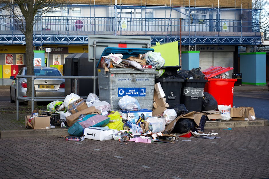 A collection of overflowing waste bins and scattered rubbish on a city sidewalk, situated near a parked silver car with a visible license plate. The bins include a large gray mixed paper and cardboard container, a black bin for general waste, a red recycling bin, and a black bin designated for recyclable packaging. The surrounding waste consists of crumpled paper, cardboard boxes, plastic bags, and packaging materials, some spilling onto the brick-paved pavement. Behind the scene, there is a building with a blue scaffolding structure and a row of shops, including a sign for a fish bar, indicating an urban commercial area. A metal safety barrier partially encloses the area, and a leafless tree is visible to the left. The scene appears to demonstrate an example of waste that would require private rubbish removal services, such as those offered by Rubbish Removal Marylebone, highlighting the need for timely collection to prevent litter build-up.