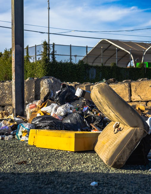 Three large black garbage bags filled with waste material are placed on the sidewalk near a black metal fence and a brick wall. The bags are made of thick plastic and appear to contain household or garden refuse, with some partially bulging and showing creases and folds on their surfaces. They are positioned in a loose cluster, with one bag leaning against the fence and the other two lying flat on the pavement. The scene is outdoors, with an area of greenery visible behind the fence, and natural light illuminating the scene, creating subtle shadows on the bags and the ground. The image conveys an outdoor rubbish collection setting, typical of private waste removal services offered by companies like Rubbish Removal Marylebone, and indicates a situation where household waste has been temporarily placed for collection, possibly as part of a local or alternative waste management process.
