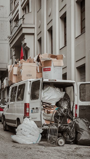 A white van parked along a narrow urban street with its rear doors open, revealing a large quantity of mixed waste and rubbish bags piled inside. Several black and white garbage bags, along with cardboard boxes and wooden pallets, are visible both inside and spilling out of the vehicle. The van is surrounded by additional rubbish bags placed on the pavement beside it, including a white bag with handwritten text. The background features multi-storey buildings with light-colored facades and windows, indicating a city centre or residential area. The scene suggests an on-site clearance operation, utilizing a private van for waste collection, typical of independent rubbish removal services such as those offered by Rubbish Removal Marylebone, especially when dealing with household or commercial waste needing alternative disposal methods.