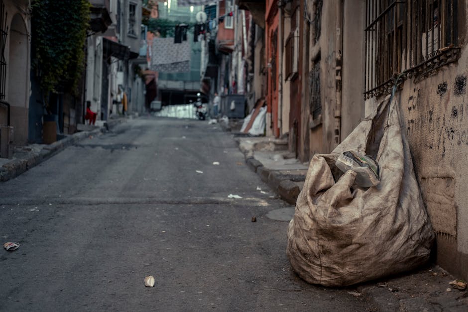 A large, dirty, beige plastic rubbish sack is placed on a narrow, uneven urban street pavement, resting against a wall with an iron barred window. The sack appears overfilled, with miscellaneous waste materials protruding from its top, including a plastic bag. Surrounding the sack, the street surface shows signs of wear, with patchy asphalt and scattered debris, such as crumpled paper and small bits of litter. In the background, an incline leads further into a residential area with multi-storey buildings, some with laundry hanging outside on makeshift lines, and others showing signs of aging lemon plasterwork. The environment has a muted, natural daylight atmosphere, highlighting the urban setting where private waste disposal methods, such as individual rubbish sacks, are typical, underscoring the context of independent collection or alternative waste handling services like those offered by Rubbish Removal Marylebone. The scene encapsulates a typical street scene that may require professional rubbish removal to prevent clutter and maintain cleanliness in the area.