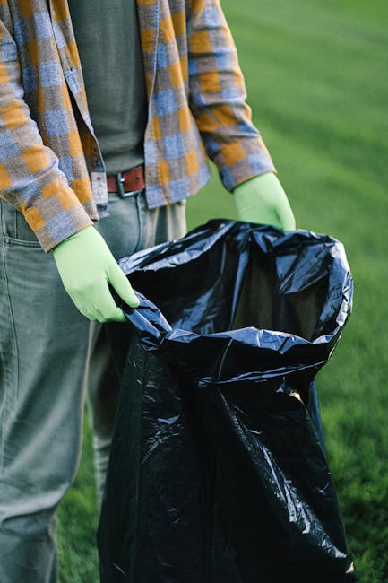 Two black plastic rubbish bags, slightly crumpled and tied at the top, are placed on a pavement near the edge of a street. The bags appear to contain household waste and are positioned close to a black metal fence with vertical bars, which divides the sidewalk from a garden or yard area with dense foliage and trees in the background. The scene is captured in monochrome, emphasizing the contrast between the dark bags and the lighter pavement surface, with some shadows cast by nearby objects and the fence. The setting suggests an informal rubbish disposal point, possibly managed by a private waste removal service such as Rubbish Removal Marylebone, rather than official municipal collection, indicating an alternative waste handling scenario within an urban environment.