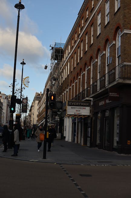 A busy urban street scene in Marylebone, featuring a row of multi-story brick buildings with large, arched windows and small balconies. The buildings’ façades are in warm brown and red tones with some white accents around the windows. On the ground level, various retail shops and businesses are visible, including a store with a black sign and white lettering, and a construction site with scaffolding and protective netting on one of the buildings. Pedestrians, dressed in casual clothing, walk along the pavement near a traffic light showing a red pedestrian signal. Streetlamps and decorative street ornaments hang overhead, and the sky is partly cloudy with patches of blue. The scene captures an active city environment consistent with an area where private waste collection services, such as those offered by Rubbish Removal Marylebone, might operate to manage rubbish and debris during property renovations or commercial activity, supporting independent waste handling in the district.