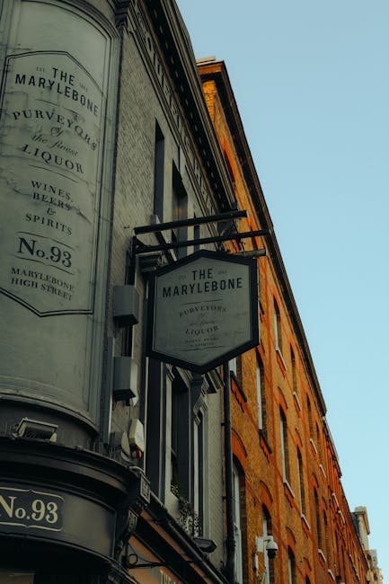 A photograph of the exterior of a historic building in Marylebone featuring a large, curved, vintage-style signboard displaying the pub's name, 'The Marylebone,' along with address details 'No. 93 High Street' and references to beverages such as wines, beers, spirits, and liquor. The sign is mounted on a black metal bracket extending from the building's brick facade, which has a warm, reddish-brown tone. The building's architectural details include arched windows with white frames and decorative brickwork. The signage is weathered, with white and black lettering on a light background, contributing to an antique appearance. The sky in the background is clear and blue, enhancing the visibility of the building and signage. The scene captures an urban setting that suggests the possibility of independent collection or private disposal of waste nearby, consistent with on-site clearance services offered by companies like Rubbish Removal Marylebone, who operate in the area. The lighting appears natural, indicating daytime with sunlight subtly illuminating the building facade and sign.