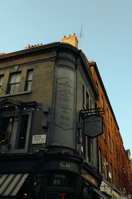 A busy urban street scene in Marylebone, featuring a row of multi-story brick buildings with large, arched windows and small balconies. The buildings’ façades are in warm brown and red tones with some white accents around the windows. On the ground level, various retail shops and businesses are visible, including a store with a black sign and white lettering, and a construction site with scaffolding and protective netting on one of the buildings. Pedestrians, dressed in casual clothing, walk along the pavement near a traffic light showing a red pedestrian signal. Streetlamps and decorative street ornaments hang overhead, and the sky is partly cloudy with patches of blue. The scene captures an active city environment consistent with an area where private waste collection services, such as those offered by Rubbish Removal Marylebone, might operate to manage rubbish and debris during property renovations or commercial activity, supporting independent waste handling in the district.