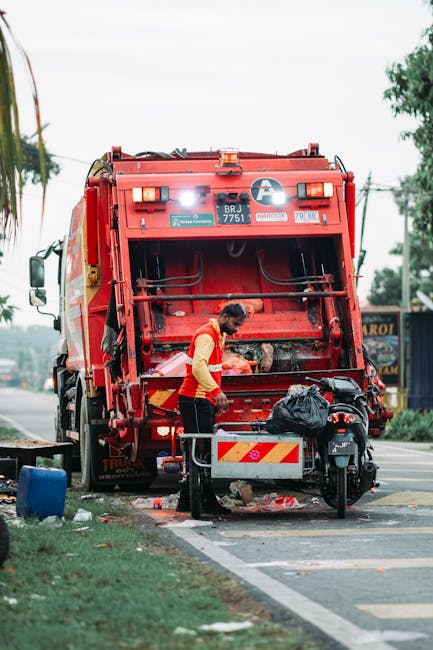 A waste collection worker wearing a high-visibility yellow and red vest is positioned behind a large red rubbish collection truck on a street, with the rear loading mechanism open and filled with black refuse bags. The worker is using a tool to manage or sort waste within the truck, which is parked at the roadside close to a curb and partial sidewalk. The truck's exterior shows identification labels, including a white circle with a blue symbol and vehicle registration details. Nearby, there is a motorbike parked adjacent to the truck, with a black bag placed on it. The environment includes a partly cloudy sky, with trees and power lines visible in the background, and a sign for a local business further along the street. This scene illustrates a typical waste collection process related to private rubbish removal services, such as those provided by Rubbish Removal Marylebone, emphasizing on-site clearance and efficient waste handling.