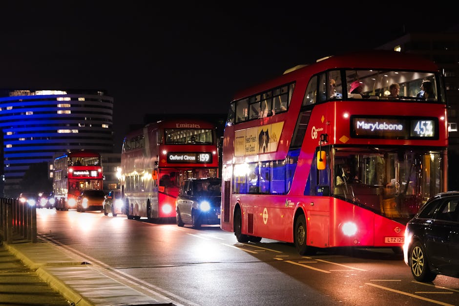 A line of three double-decker buses illuminated by streetlights at night on a city street. The front bus, displaying route number 453 headed to Marylebone, is red with a black front window frame and visible interior lighting showing passengers seated inside. Behind it, a second red double-decker bus with route number 159 to Oxford Circus is also illuminated, and further back, a third red bus can be partially seen. To the right of the buses, a few parked cars are visible along a curbside, with some headlights casting additional light onto the scene. The background features dark night sky and modern buildings, including a distinctive structure with curved horizontal lines lit in blue. The scene conveys an urban environment with active public transportation, relevant in the context of private or alternative waste management and rubbish removal services by companies such as Rubbish Removal Marylebone.