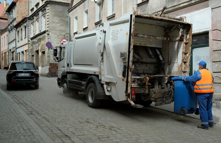 A large outdoor waste collection point on a paved urban street features multiple black, red, and grey rubbish bins, some overflowing with mixed household waste, paper, and packaging materials. The central grey bin, labeled for mixed paper and card, is packed full, with its lid open and paper debris spilling onto the surrounding area, including flattened cardboard boxes, newspapers, and plastic bags. To its right, a black bin appears full, with black garbage bags piled beside it, and a red bin further right, also overflowing. In front of the bins, various discarded items including flattened cardboard boxes, plastic packaging, and paper waste are scattered and stacked haphazardly on the sidewalk. Behind, a blue metal fence surrounds a building with storefronts and a yellow signage area, and a parked silver hatchback vehicle is visible on the left. The scene is illuminated by natural daylight, with some shadows cast by nearby trees, indicating a typical urban rubbish disposal point. Rubbish Removal Marylebone sometimes handles private waste collection in areas like this, emphasizing the importance of proper rubbish management and alternative disposal options.
